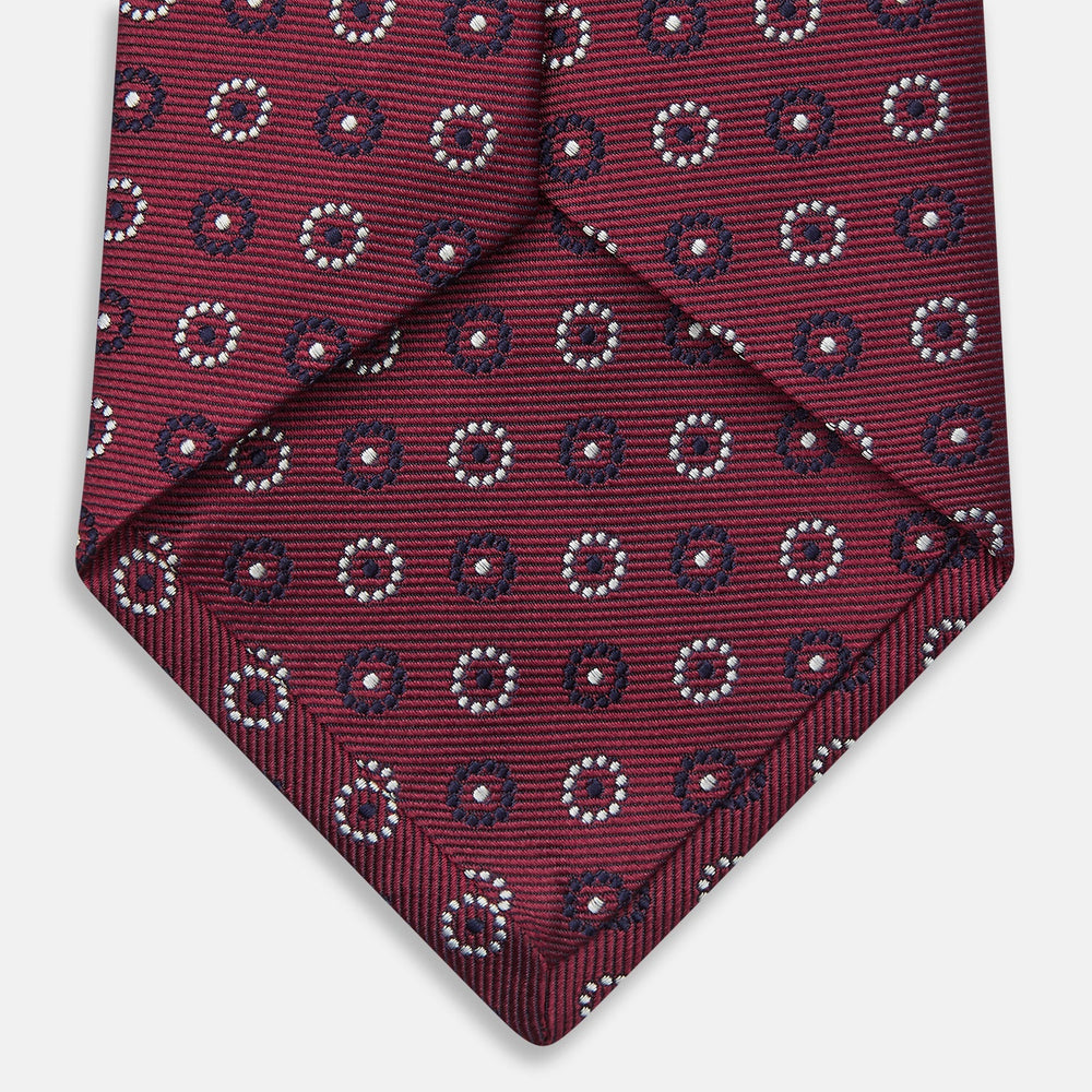The Red Spheric Emblem Tie by Turnbull & Asser is a rolled-up maroon silk tie with a jacquard pattern of small white and purple circular dots, shown against a white background.