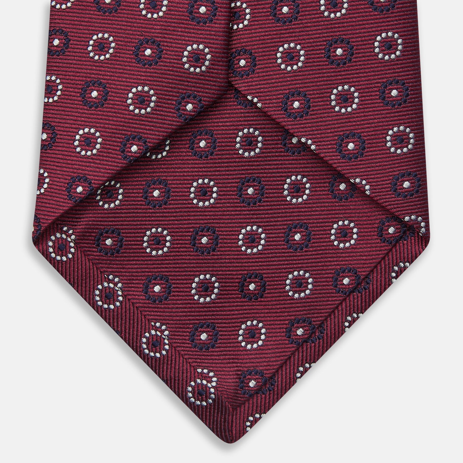 View 4 of product The Red Spheric Emblem Tie by Turnbull & Asser is a rolled-up maroon silk tie with a jacquard pattern of small white and purple circular dots, shown against a white background.