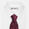 View 2 of product The Red Spheric Emblem Tie by Turnbull & Asser is a rolled-up maroon silk tie with a jacquard pattern of small white and purple circular dots, shown against a white background.