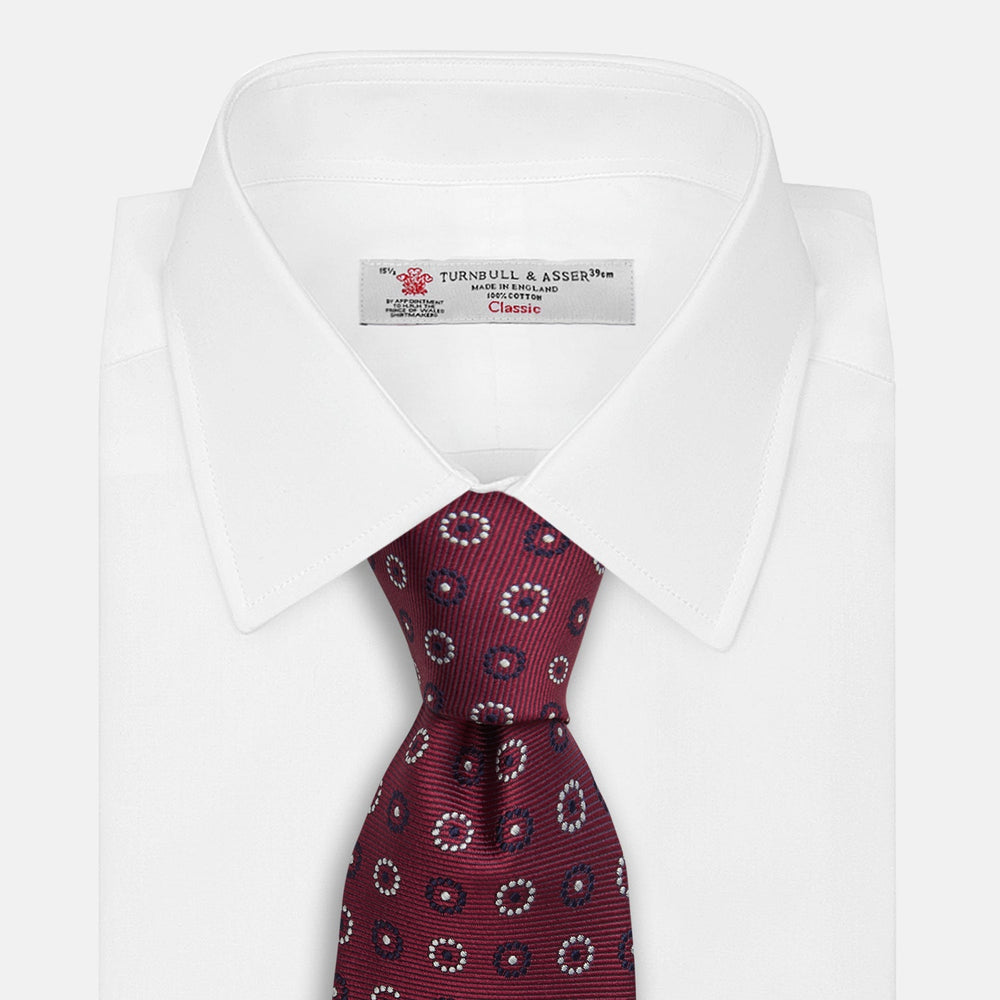 The Red Spheric Emblem Tie by Turnbull & Asser is a rolled-up maroon silk tie with a jacquard pattern of small white and purple circular dots, shown against a white background.