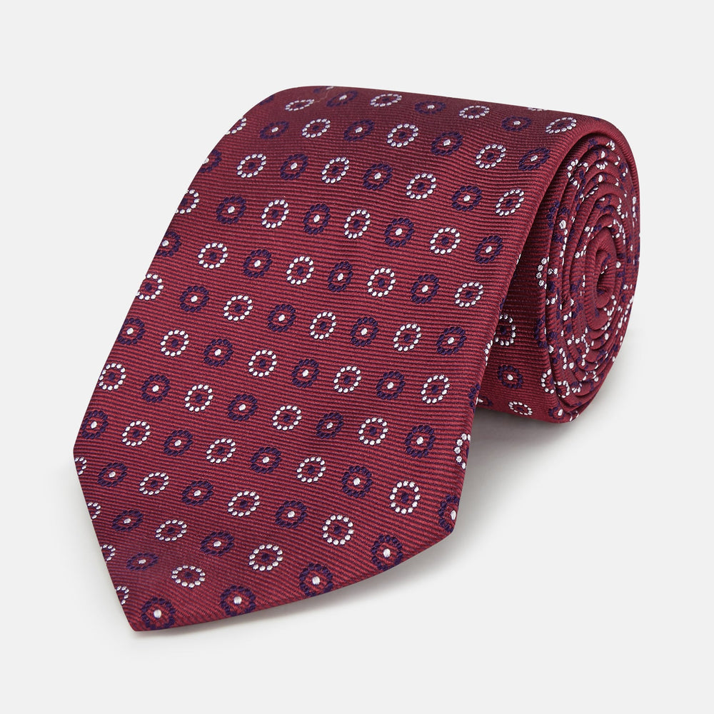 The Red Spheric Emblem Tie by Turnbull & Asser is a rolled-up maroon silk tie with a jacquard pattern of small white and purple circular dots, shown against a white background.