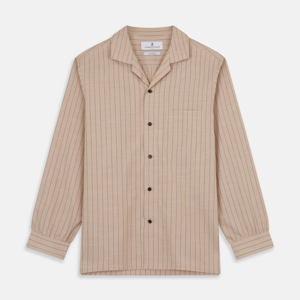 An older man in a light hat, Turnbull & Asser Beige Stripe Cotton Holiday Fit Shirt with Revere Collar & Single Button Cuff, and white pants leans casually against a tall concrete pillar outdoors, greenery blurred behind him.
