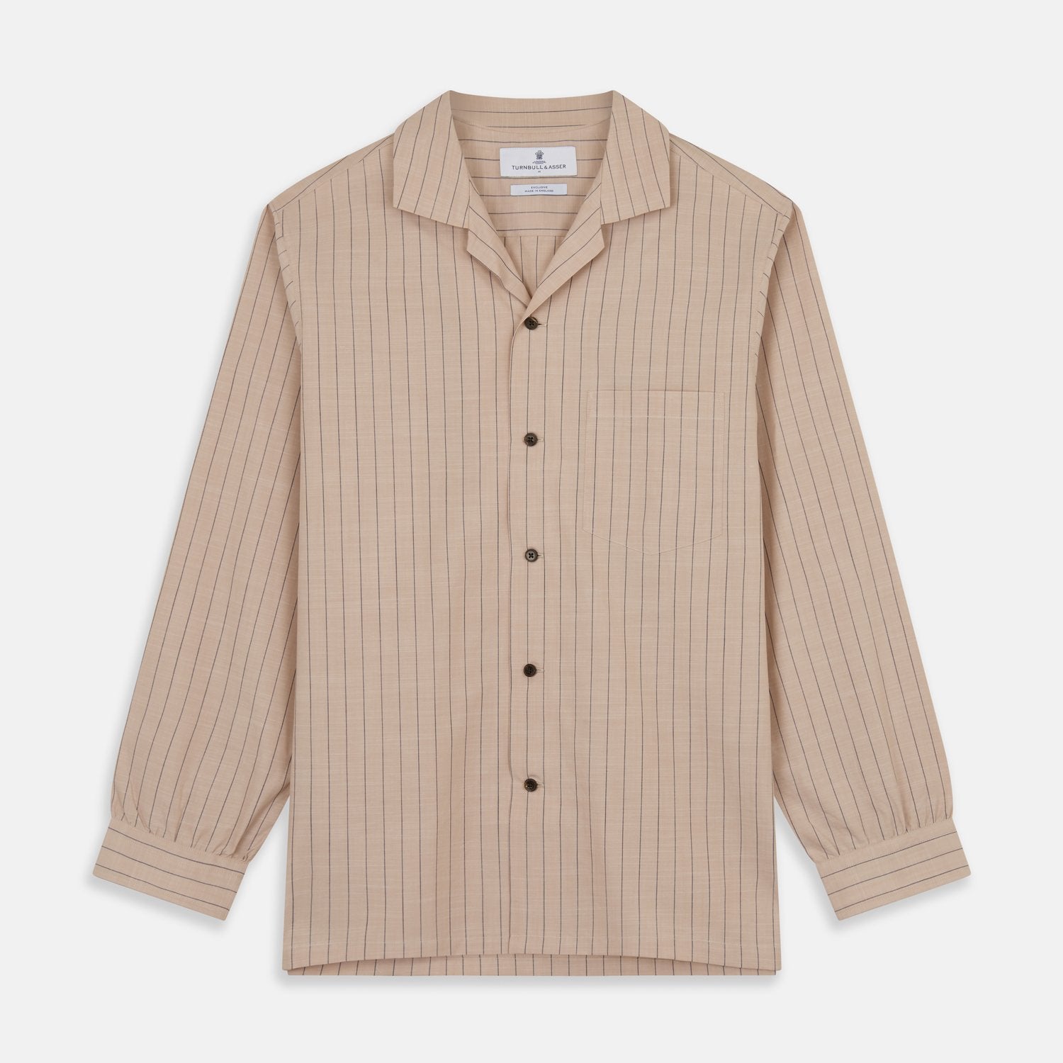 View 2 of product An older man in a light hat, Turnbull & Asser Beige Stripe Cotton Holiday Fit Shirt with Revere Collar & Single Button Cuff, and white pants leans casually against a tall concrete pillar outdoors, greenery blurred behind him.