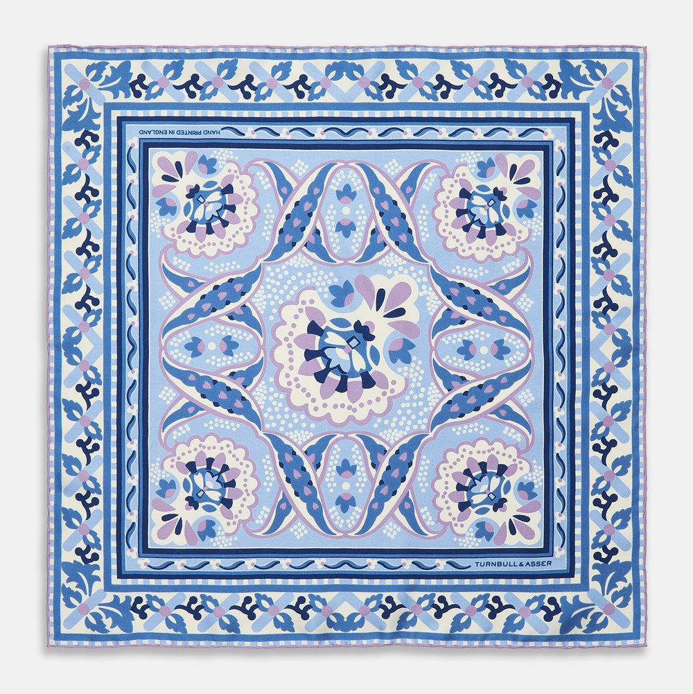 The Pale Blue and Lilac Baroque Silk Pocket Square by Turnbull & Asser features intricate geometric and floral patterns in blue, lilac, and white with a floral border and the brand name printed in one corner.