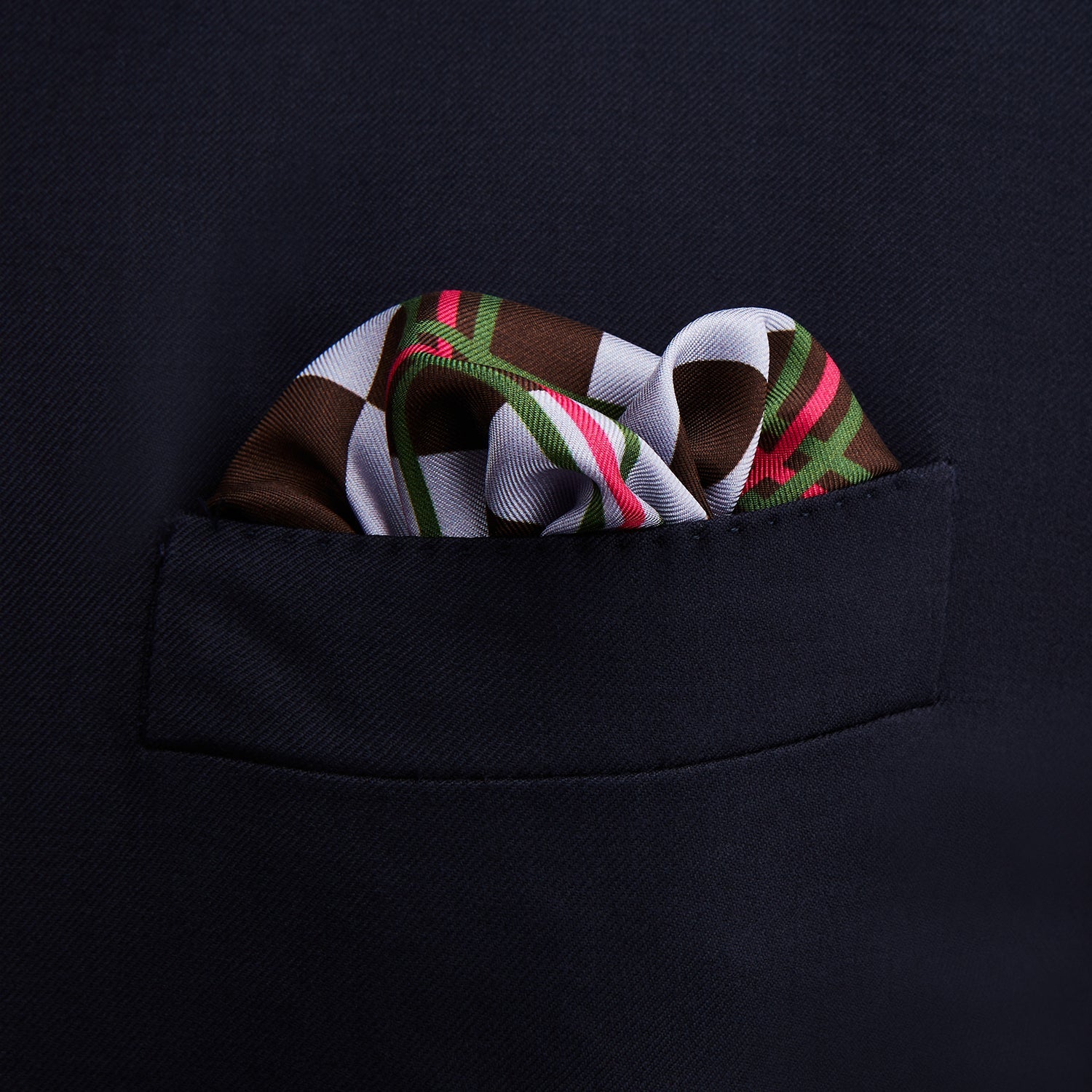 View 3 of product The Turnbull & Asser Green and Red Checked Chess Board Silk Pocket Square features brown, green, red, and white stripes with a thin green border and the brand's name in the bottom right corner.