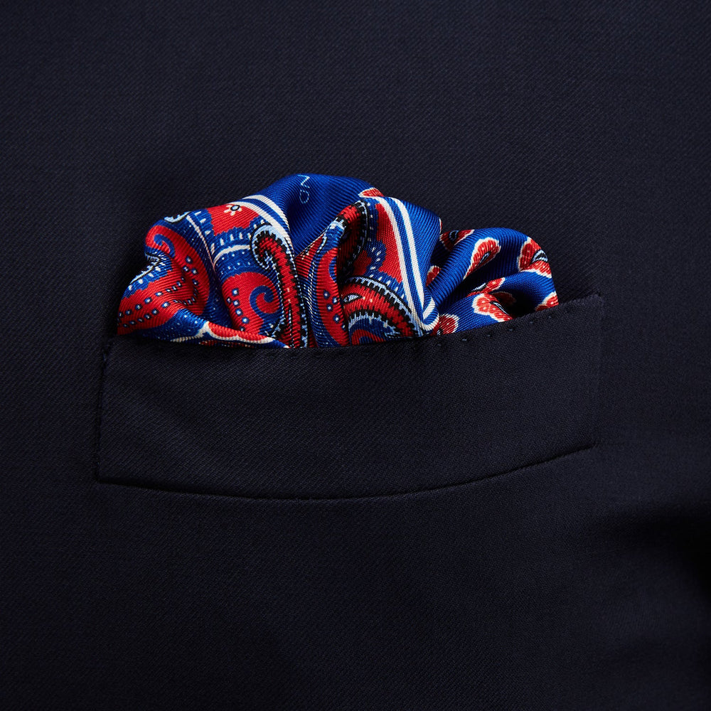 The Blue and Red Paisley Tiles Silk Pocket Square by Turnbull & Asser features a blue background, red and white paisley border, central repeating circular patterns, and “Turnbull & Asser” in one corner. Made in England.