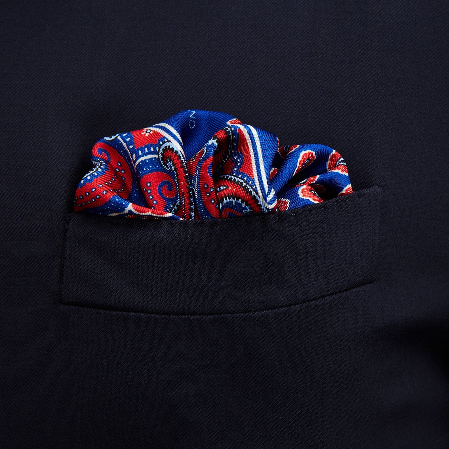 View 3 of product The Blue and Red Paisley Tiles Silk Pocket Square by Turnbull & Asser features a blue background, red and white paisley border, central repeating circular patterns, and “Turnbull & Asser” in one corner. Made in England.
