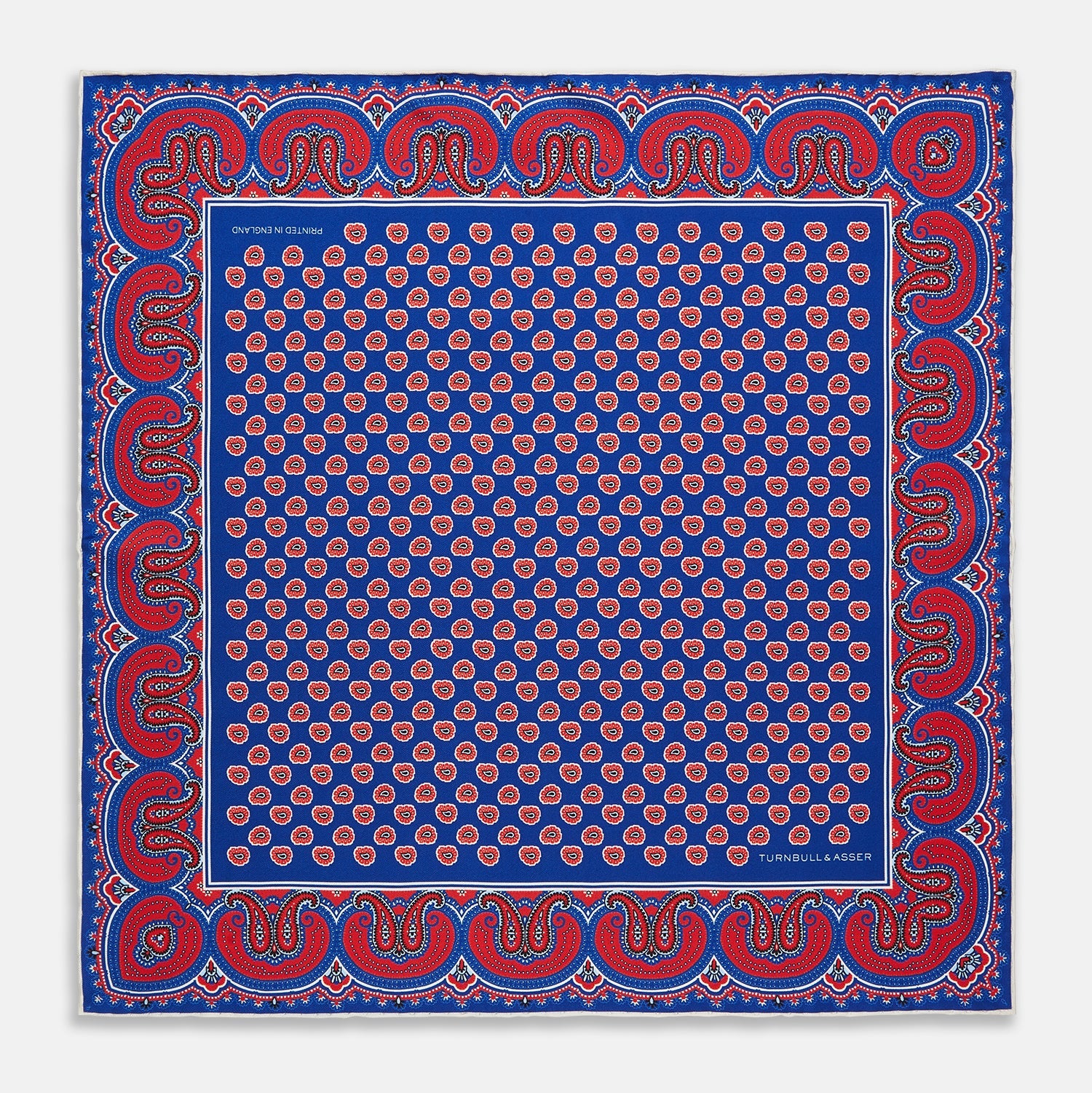 View 1 of product The Blue and Red Paisley Tiles Silk Pocket Square by Turnbull & Asser features a blue background, red and white paisley border, central repeating circular patterns, and “Turnbull & Asser” in one corner. Made in England.