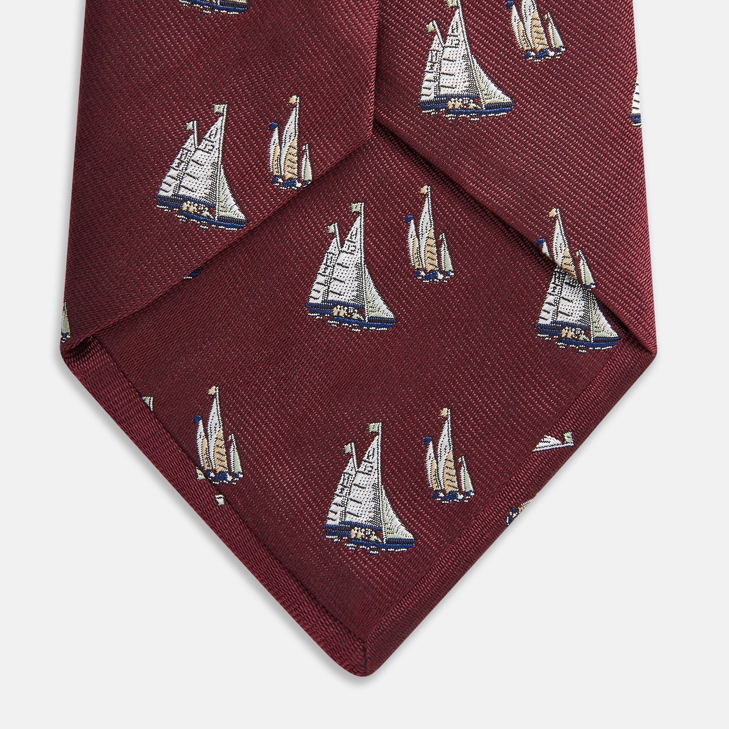 View 3 of product The Turnbull & Asser Burgundy Racing Yachts Silk Tie features a nautical sailboat pattern in white, blue, and brown on burgundy silk, shown rolled up against a plain white background.