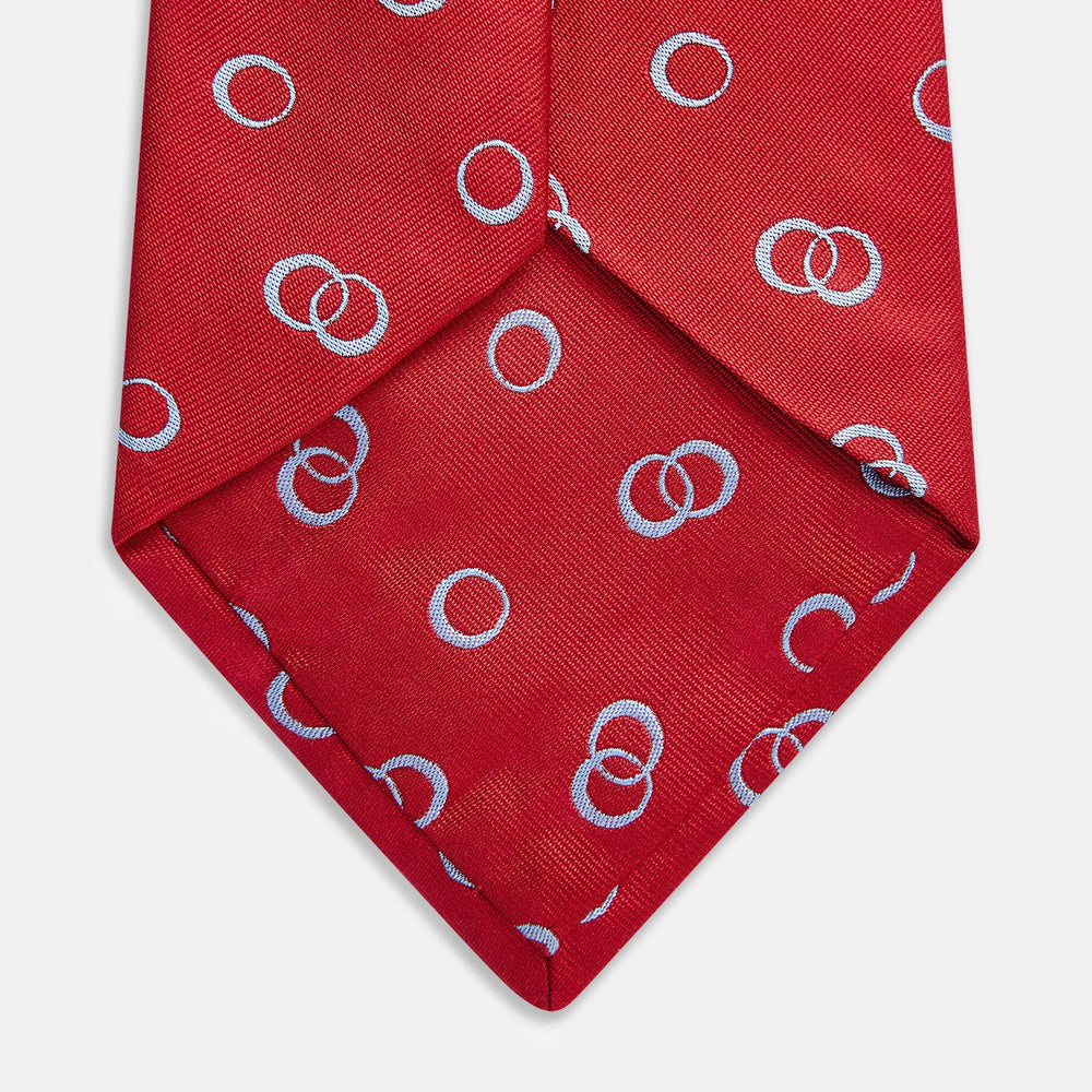 The Turnbull & Asser Red Geometric Rings Silk Tie, made in England, features a rolled-up design with small overlapping light blue circles on red silk and is displayed against a plain white background.