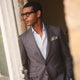 A man in glasses and a Turnbull & Asser Brown & Blue Check Merino Barrington Blazer, detailed with a colorful pocket square, stands thoughtfully indoors by patterned curtains, gazing out the window.