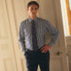 A person in a Turnbull & Asser Navy & White Stripe Weekend Fit Shirt with Derby Collar and Single Button Cuffs, paired with a paisley tie and dark pants, stands indoors leaning against a cream paneled wall.