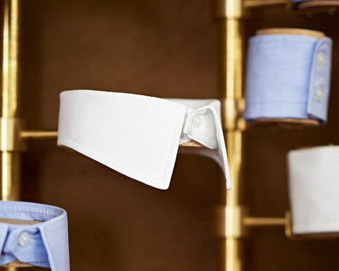 A close-up of a white detachable shirt collar displayed on a gold rack, surrounded by rolled-up shirt cuffs in pale blue and white fabrics.