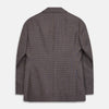 View 5 of product A man in glasses and a Turnbull & Asser Brown & Blue Check Merino Barrington Blazer, detailed with a colorful pocket square, stands thoughtfully indoors by patterned curtains, gazing out the window.