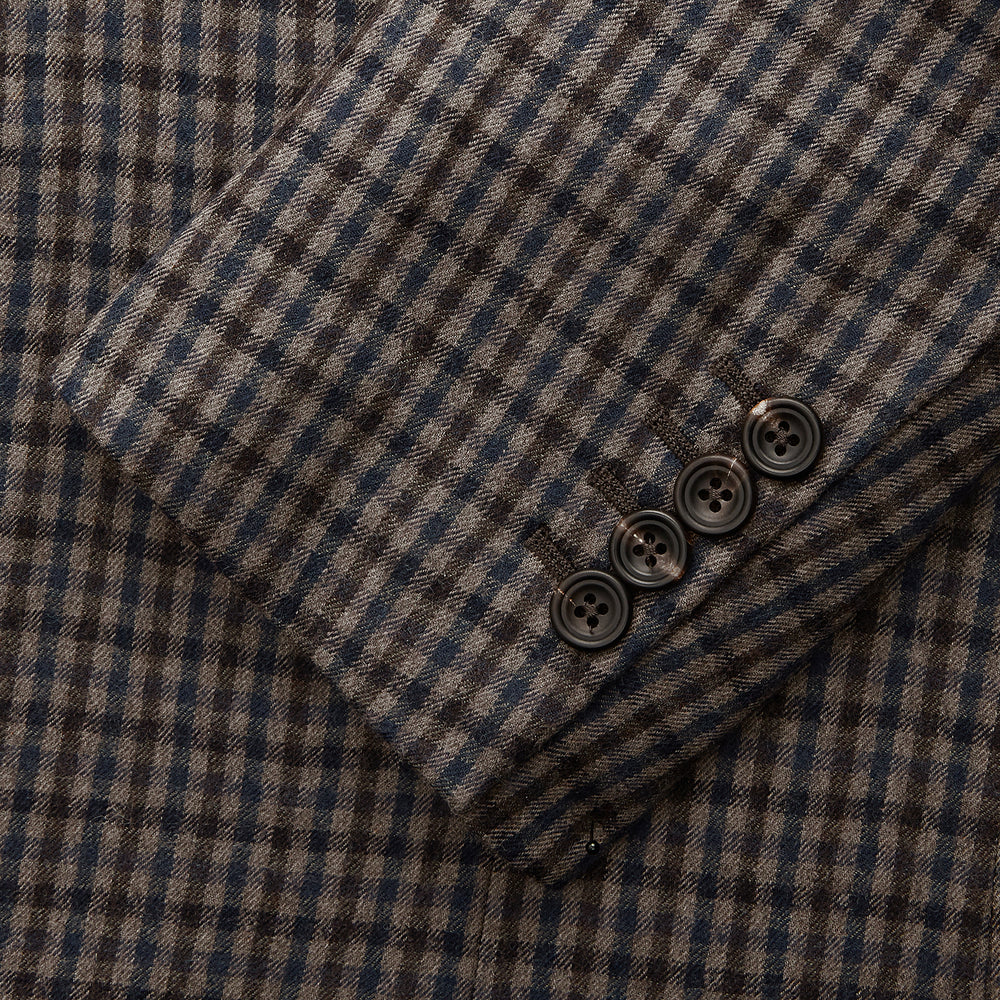 A man in glasses and a Turnbull & Asser Brown & Blue Check Merino Barrington Blazer, detailed with a colorful pocket square, stands thoughtfully indoors by patterned curtains, gazing out the window.