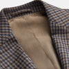 View 3 of product A man in glasses and a Turnbull & Asser Brown & Blue Check Merino Barrington Blazer, detailed with a colorful pocket square, stands thoughtfully indoors by patterned curtains, gazing out the window.