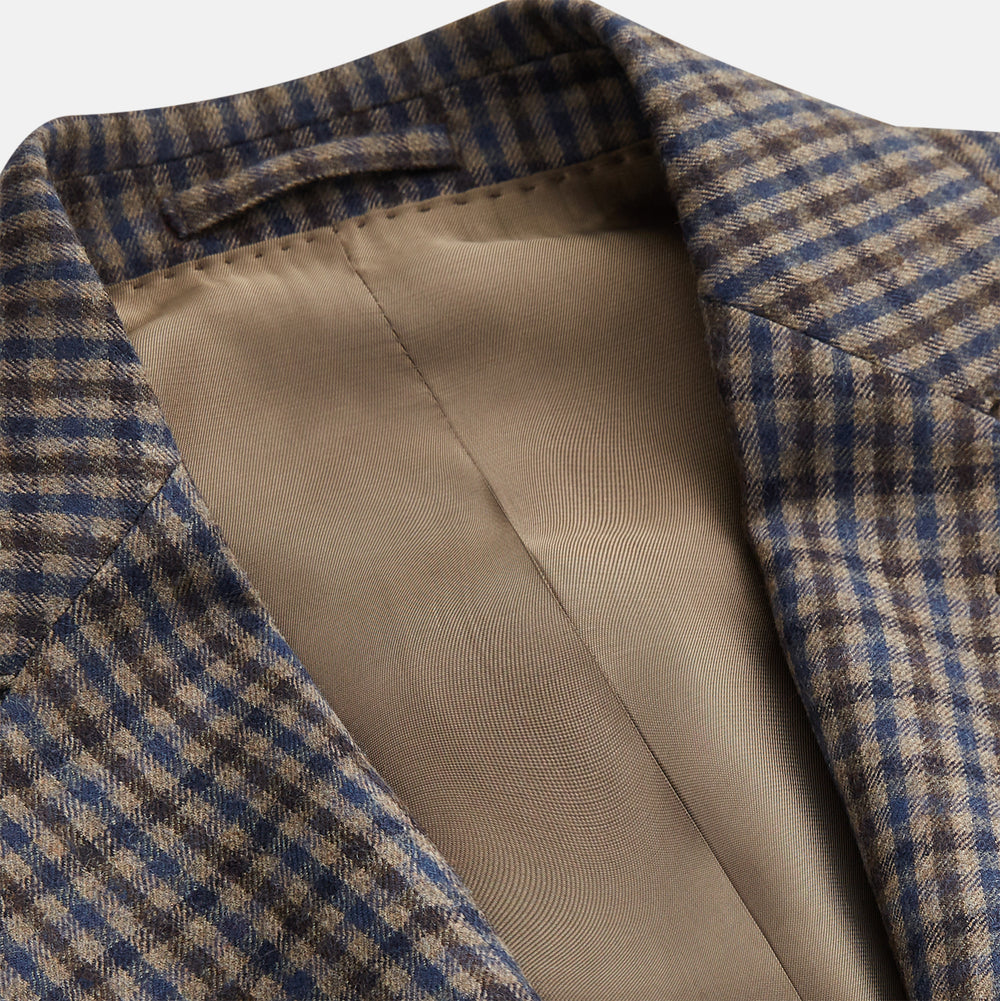 A man in glasses and a Turnbull & Asser Brown & Blue Check Merino Barrington Blazer, detailed with a colorful pocket square, stands thoughtfully indoors by patterned curtains, gazing out the window.