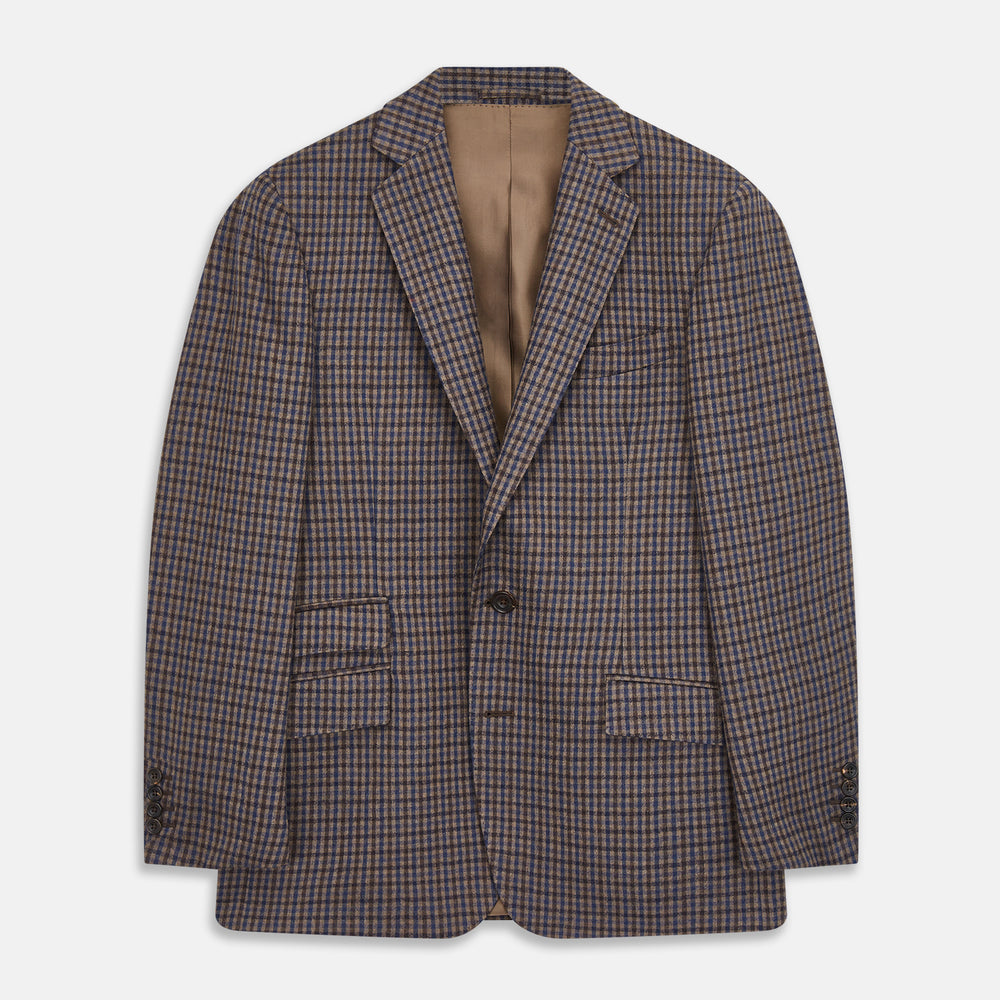 A man in glasses and a Turnbull & Asser Brown & Blue Check Merino Barrington Blazer, detailed with a colorful pocket square, stands thoughtfully indoors by patterned curtains, gazing out the window.
