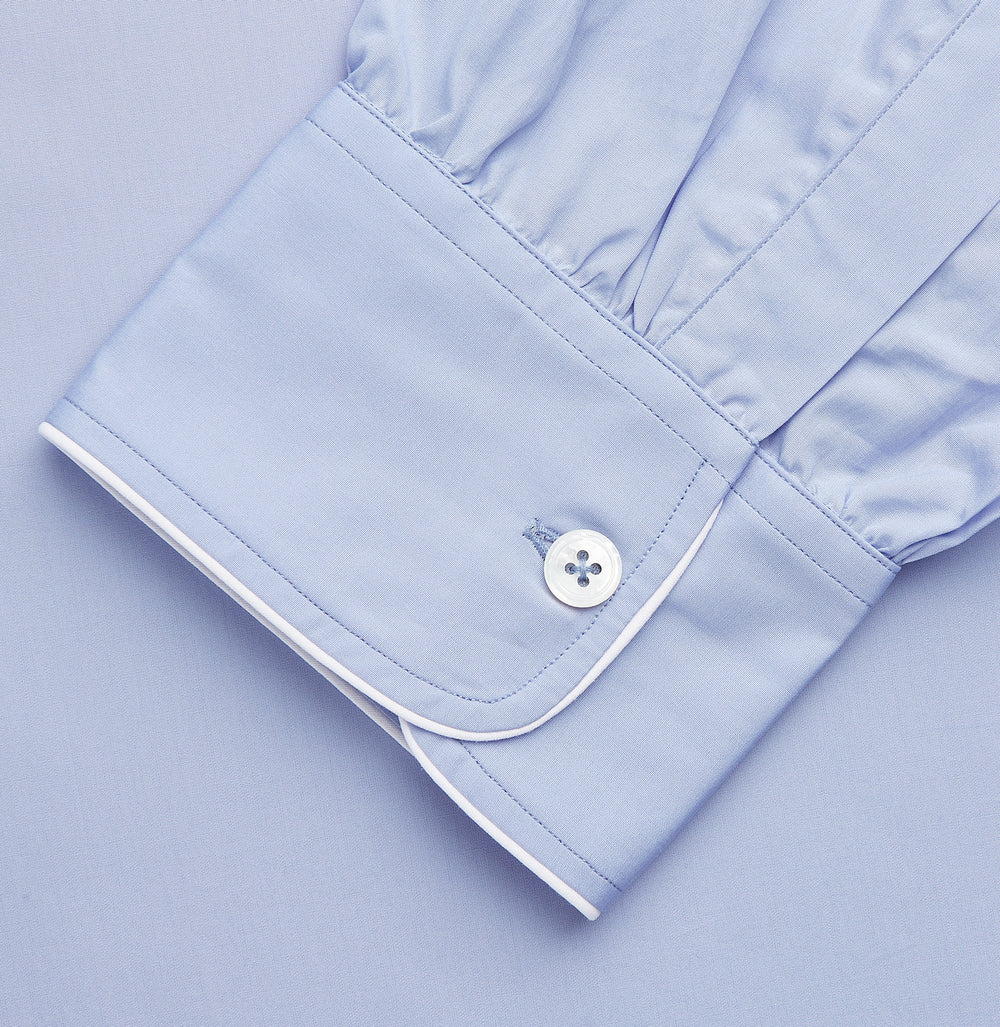 The Turnbull & Asser Blue Piped Sea Island Quality Cotton Nightshirt features light blue sea island cotton, a collared neckline with white piping, a front pocket, partial button placket, and a curved high-low hem for classic nightshirt style.