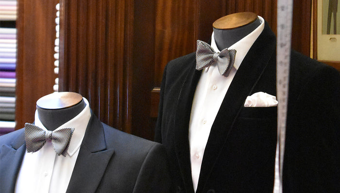 Two mannequins dressed in formal black tuxedos with white shirts and patterned bow ties are displayed in a wood-paneled store, with colorful fabrics visible in the background.
