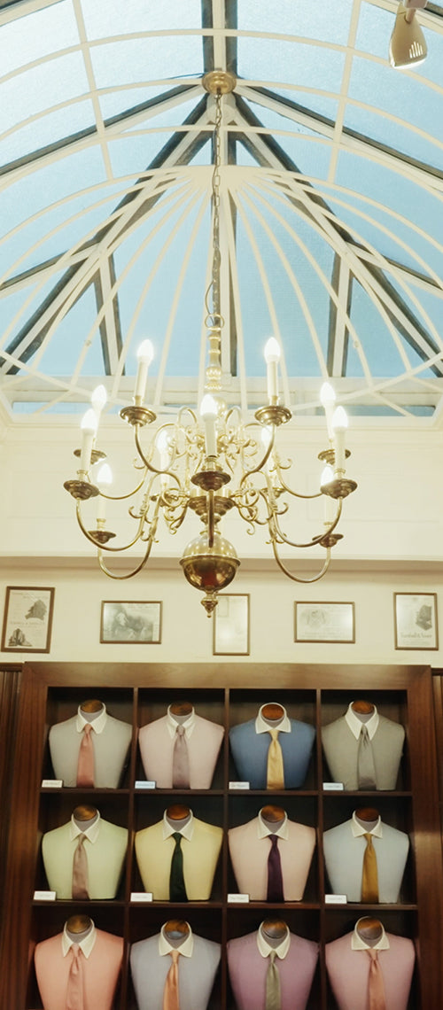 A gold chandelier hangs from a glass ceiling above a display of mannequin busts in colorful shirts and ties, arranged in wooden shelves. Framed artwork decorates the wall above the display.