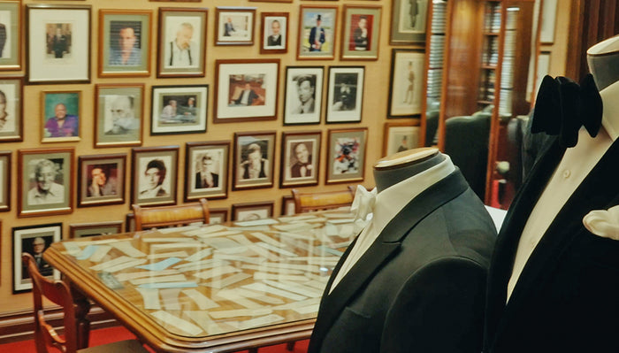 A room with walls covered in framed photographs, a glass-top table with wooden chairs, and two headless mannequins dressed in tuxedos in the foreground.