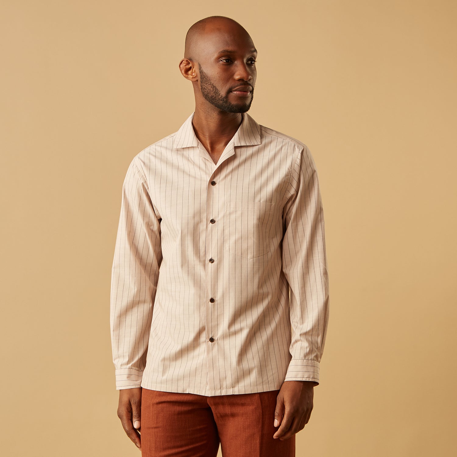 View 8 of product An older man in a light hat, Turnbull & Asser Beige Stripe Cotton Holiday Fit Shirt with Revere Collar & Single Button Cuff, and white pants leans casually against a tall concrete pillar outdoors, greenery blurred behind him.