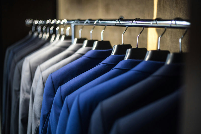 Turnbull & Asser Cloth Price suits in blue and gray hang neatly on a metal rail, displayed in a dimly lit closet or store.