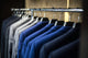 Turnbull & Asser Cloth Price suits in blue and gray hang neatly on a metal rail, displayed in a dimly lit closet or store.