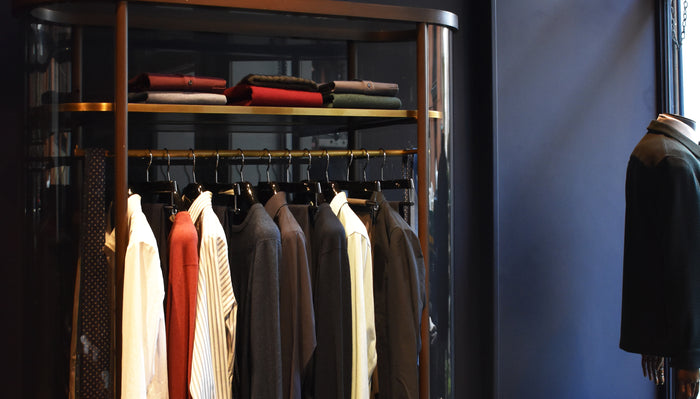 A clothing rack displays various shirts, jackets, and coats neatly hung on hangers below folded sweaters, with a mannequin in a jacket partially visible on the right against a dark blue wall.