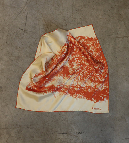 A cream-colored scarf with an orange abstract tree design is spread out on a concrete surface. The name DANIEL appears in orange text in one corner of the scarf.