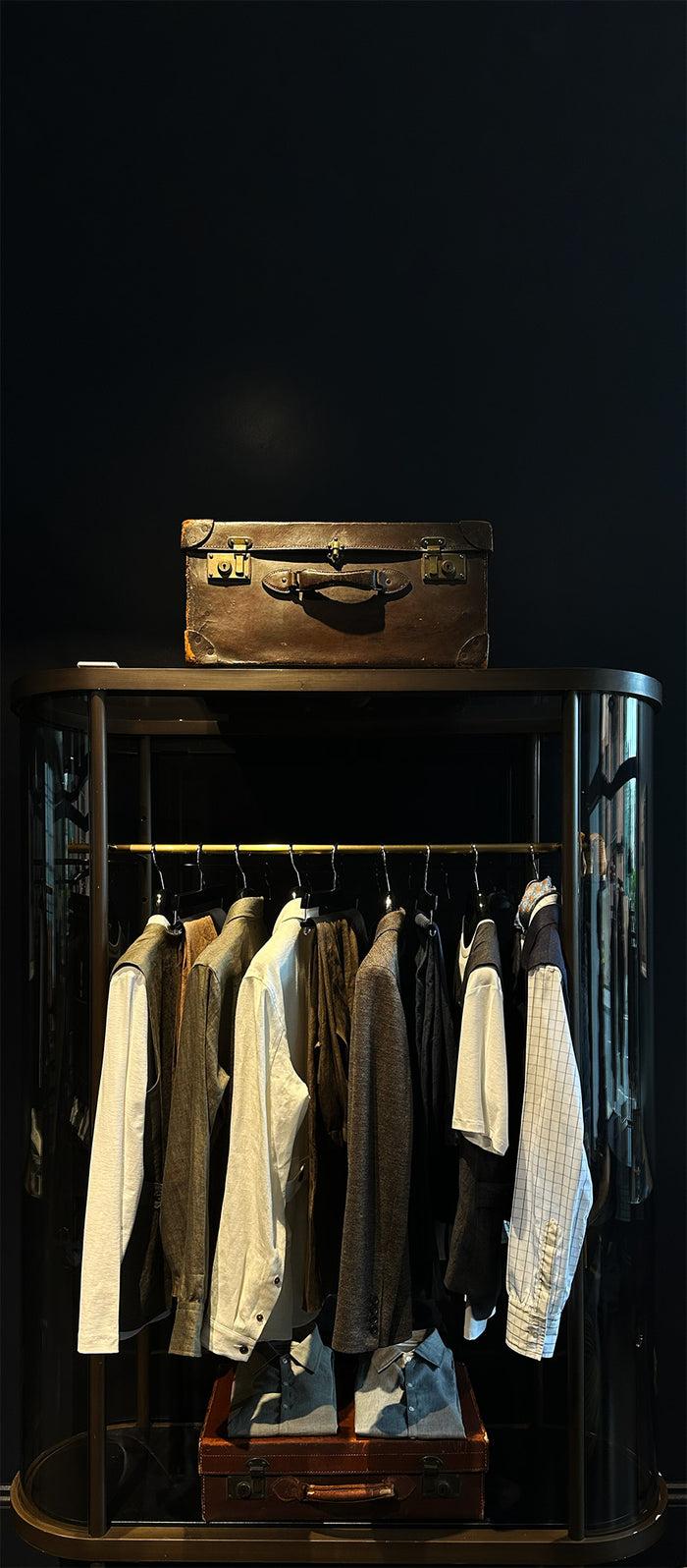 A wardrobe with several mens shirts and jackets hanging on a rail, a pair of jeans below, and a vintage brown suitcase on top, all set against a dark background.