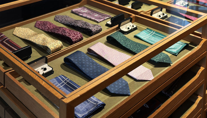 A display case with neatly arranged patterned neckties and cufflinks, set on a green felt surface inside a wooden frame. The ties are in various colors and designs, organized in rows.