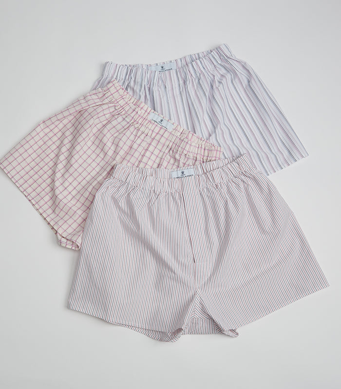 Three pairs of mens boxers in pastel colors, featuring different patterns: pink plaid, blue vertical stripes, and red vertical stripes, laid flat on a white background.