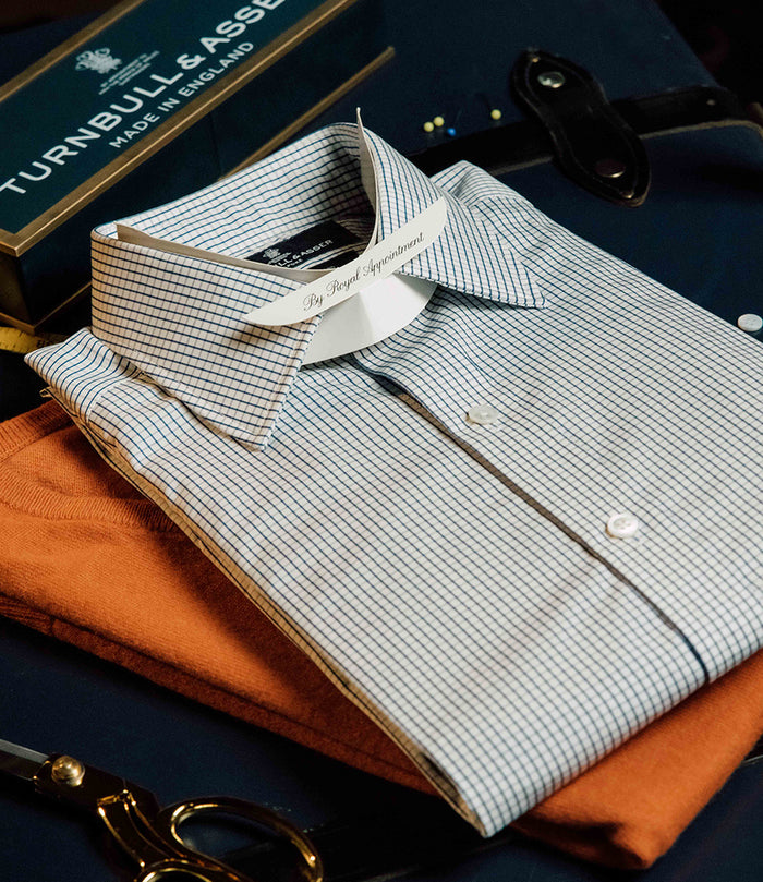 A neatly folded checkered dress shirt with a collar tag reading “By Royal Appointment” rests on top of an orange sweater. Nearby are a pair of scissors, a leather strap, and a Turnbull & Asser box.