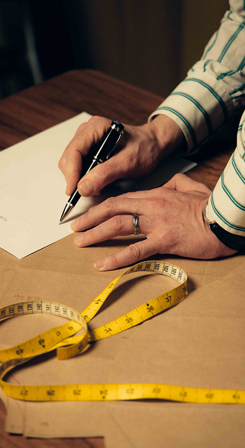 A person wearing a striped shirt writes on a piece of paper with a black pen. A yellow measuring tape is coiled on a wooden table beside the hands, one of which wears a silver ring.