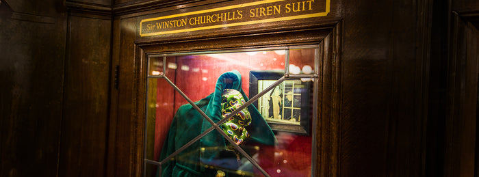 A display case with a green suit and a gas mask, labeled Sir Winston Churchills Siren Suit, set against a red background with wood paneling and a small framed photo inside the case.