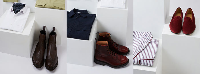 A display of mens clothing and shoes on white platforms, including folded shirts, dark brown boots, red leather boots, red velvet loafers, and striped pajamas.