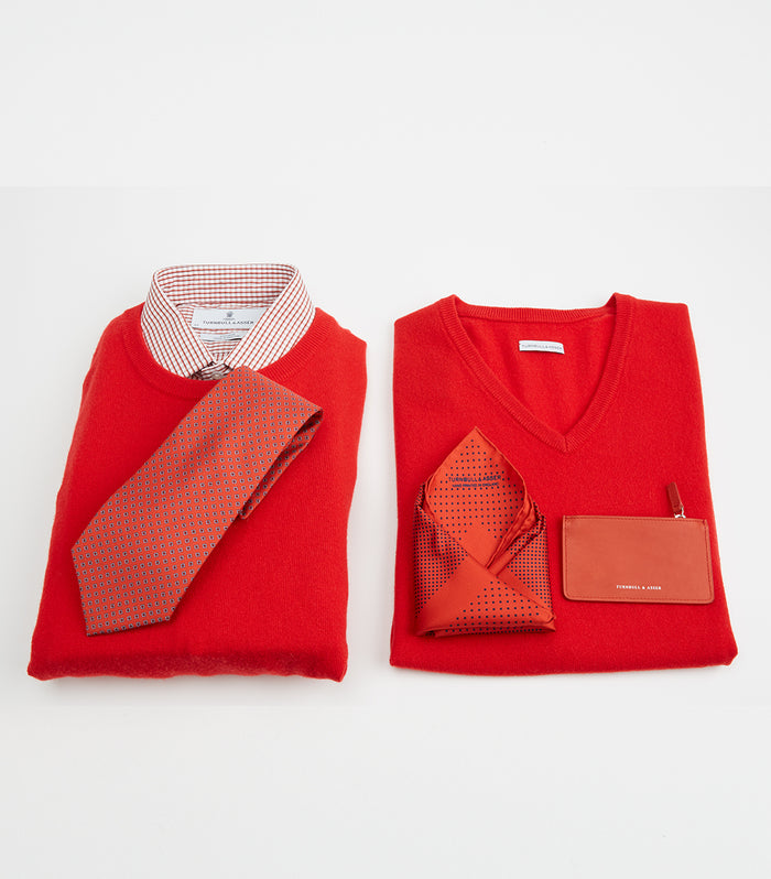 Two neatly folded red sweaters are displayed on a white background. The left sweater has a checkered shirt and a patterned tie, while the right sweater features a pocket square and a small red cardholder in the pocket.