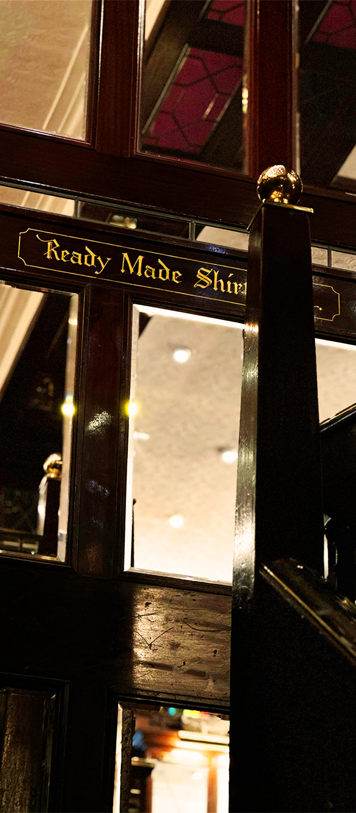 A dark wooden frame with a gold sign reading Ready Made Shi in elegant lettering, illuminated by warm indoor lighting. The background shows a ceiling with recessed lights and stained-glass panels.