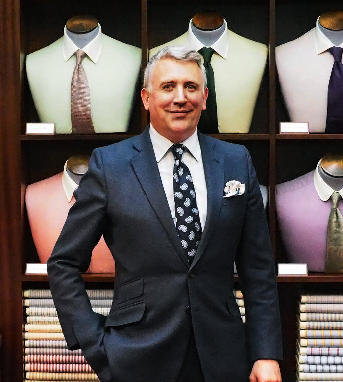 A man in a dark suit and patterned tie stands in front of a display of dress shirts and ties in various colors, with stacks of folded fabrics below. He is smiling and has one hand in his pocket.