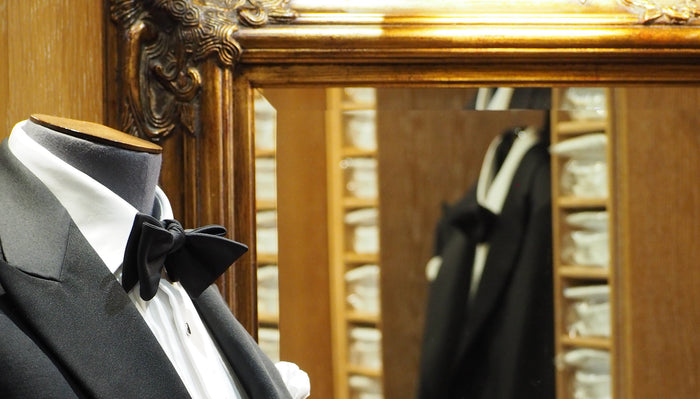 A mannequin dressed in a black tuxedo with a white shirt and black bow tie stands in front of a large ornate mirror, reflecting rows of neatly stacked shirts in a wooden display.