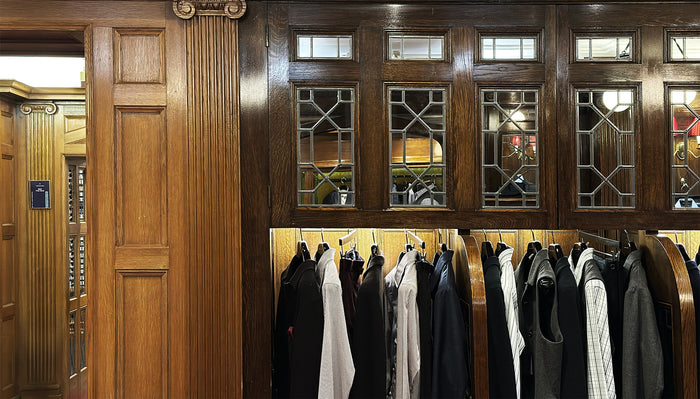 A wooden-paneled room with glass windows above several racks of formal clothing, including shirts, jackets, and vests, neatly hanging on hangers. Warm lighting creates a classic, elegant atmosphere.