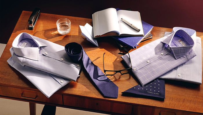 A wooden desk with two folded dress shirts, two navy ties, a pocket square, a pair of glasses, a glass of water, a stapler, an open notebook with a pen, and stacked blue notebooks.