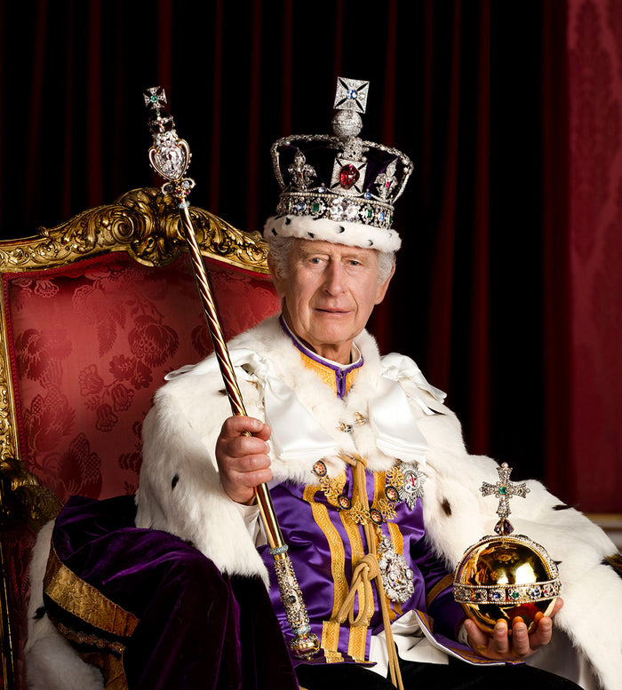 An older man in elaborate royal regalia, including a jeweled crown, fur-trimmed cloak, and ornate medals, sits on a gold-trimmed red throne, holding a ceremonial scepter and an orb against a red curtain backdrop.