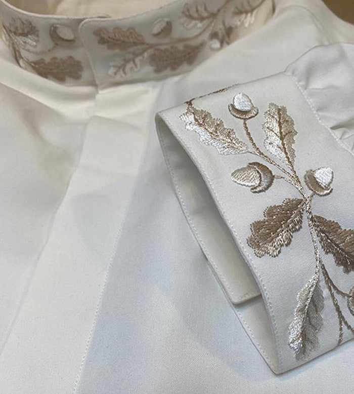 Close-up of a white shirt with intricate beige and ivory embroidery of acorns and oak leaves on the collar and cuff. The fabric appears smooth and slightly shiny.