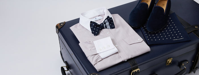 A folded dress shirt with a navy polka dot bow tie, navy loafers, and a navy polka dot pocket square are neatly arranged on top of a closed dark blue suitcase.