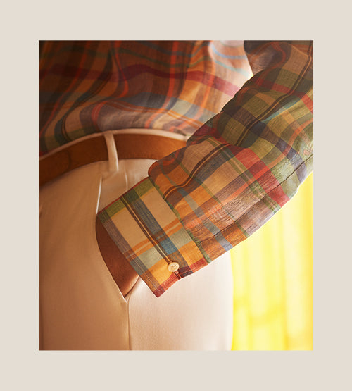 A close-up of a person wearing a colorful plaid long-sleeve shirt tucked into high-waisted, light-colored trousers with a brown belt. The image focuses on the shirt sleeve and waist area.