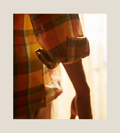 A close-up of a person’s arm wearing a colorful plaid shirt with rolled-up sleeves, bathed in warm sunlight. The background is softly blurred.