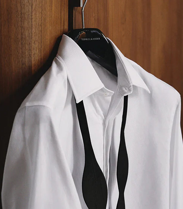 A white dress shirt hangs on a black hanger against a wooden background, with a black bow tie draped loosely around the collar.